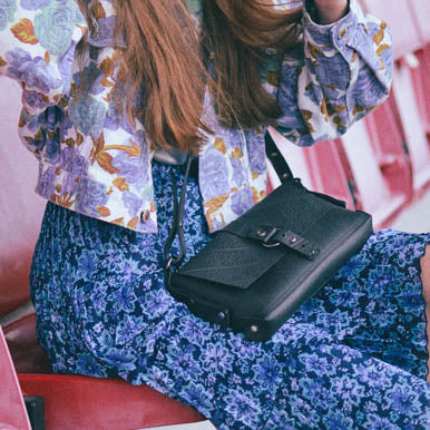 Black vegan handbag from Paguro Upcycle worn by woman with floral outfit sitting on red seat