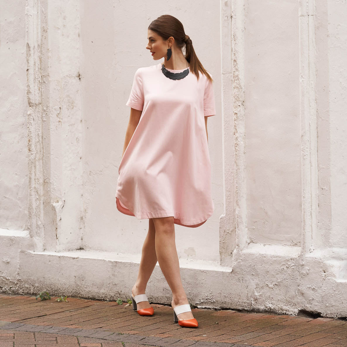 Woman wearing a pink dress and an upcycled feather necklace by Paguro Upcycle standing against a white wall
